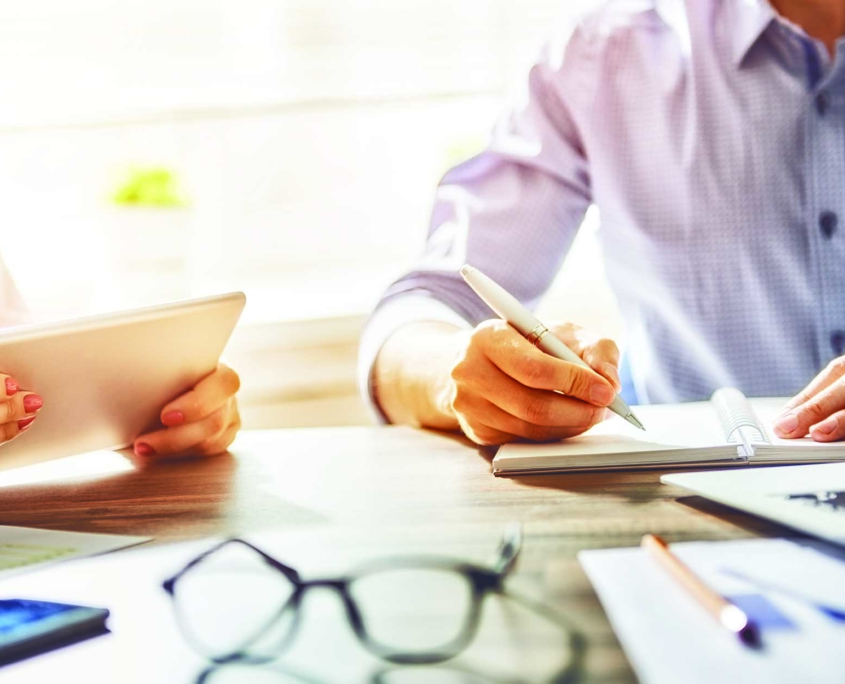 Close up view of two workers sitting at a table using a tablet and notepad