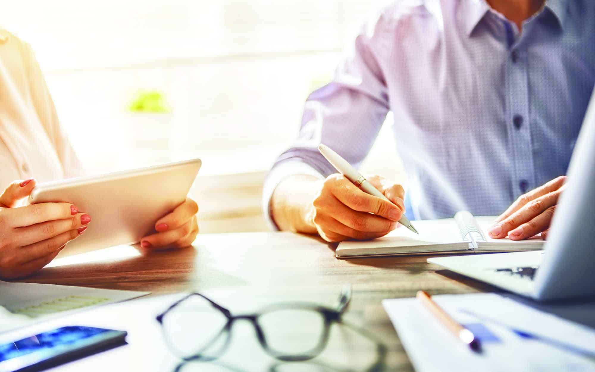 Close up view of two workers sitting at a table using a tablet and notepad