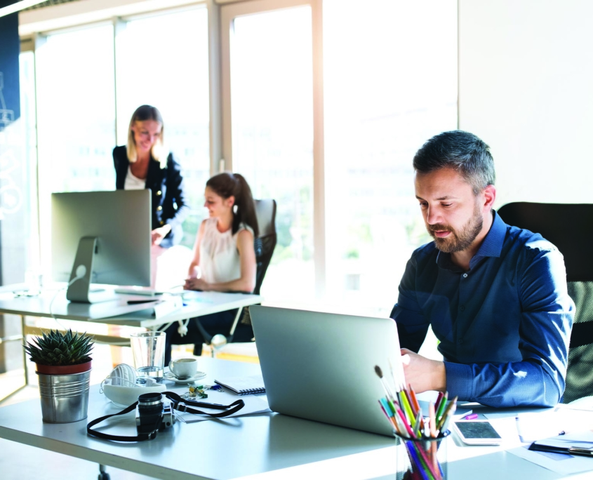 Employee looking at laptop screen behind a desk with two coworkers chatting in the background