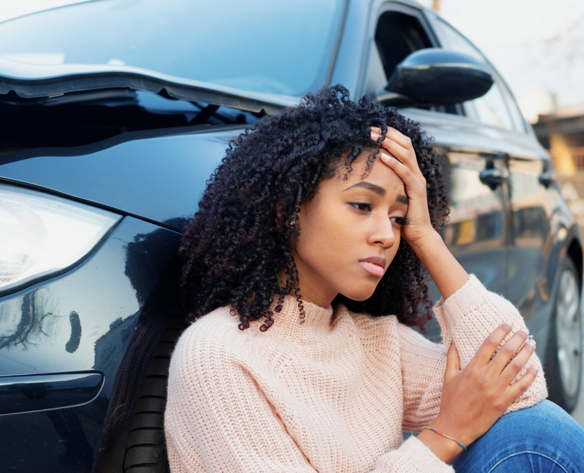 woman sits on ground and leans against damaged car after accident