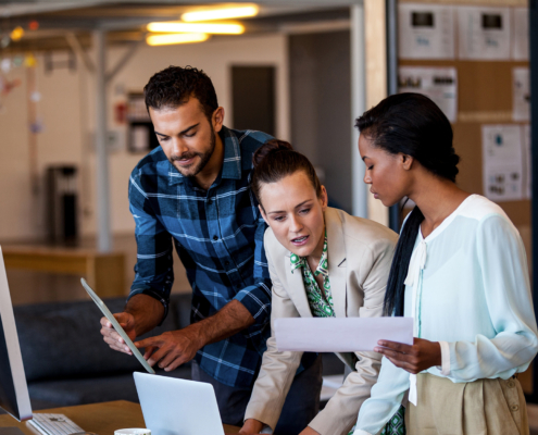 three workers in an office stand in front of desk and review documents