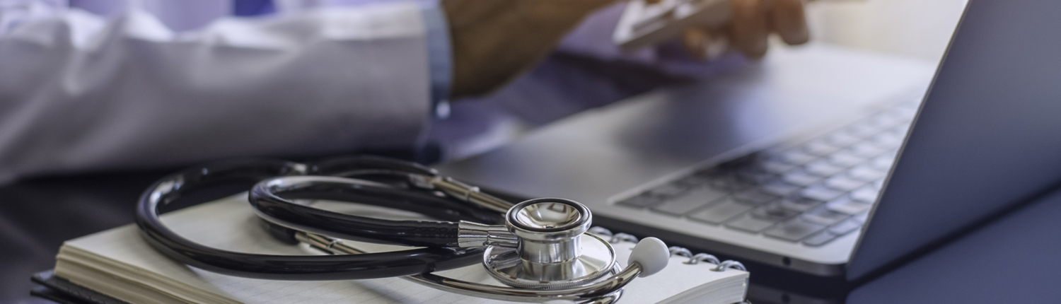 Close up of doctor using calculator in front of a laptop with notebook and stethoscope sitting nearby