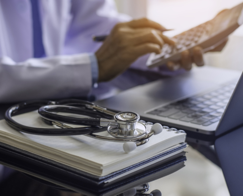 Close up of doctor using calculator in front of a laptop with notebook and stethoscope sitting nearby