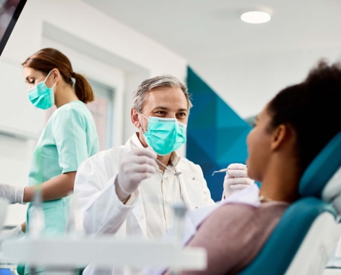 Smiling dentist with face mask talking to Black woman during dental procedure at dental clinic_